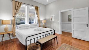 Un dormitorio con cama, mesitas de noche, un banco, una ventana con cortinas, una puerta y vistas al exterior.