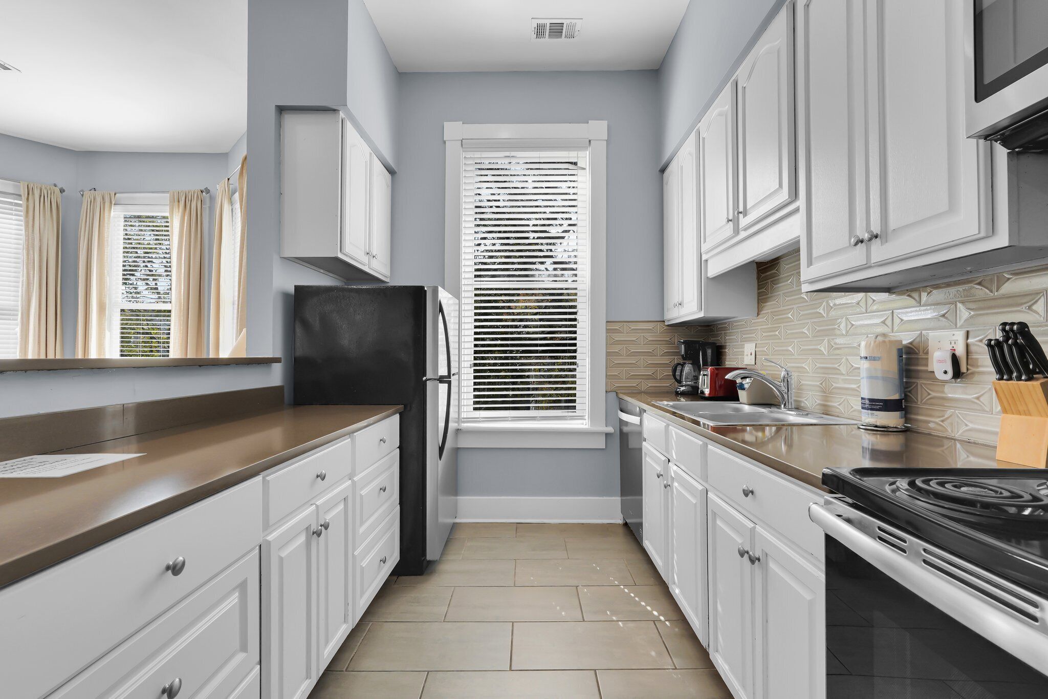 A modern kitchen with white cabinets, a black refrigerator, a window with blinds, and a tiled backsplash.