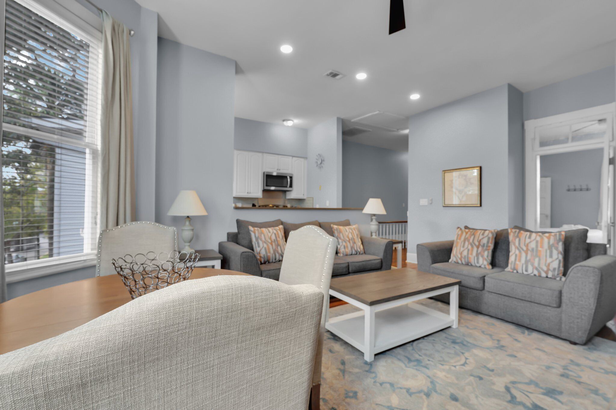 A modern living room with a grey sofa, a coffee table, and a dining table. There is a kitchen area in the background with cabinets and a microwave.