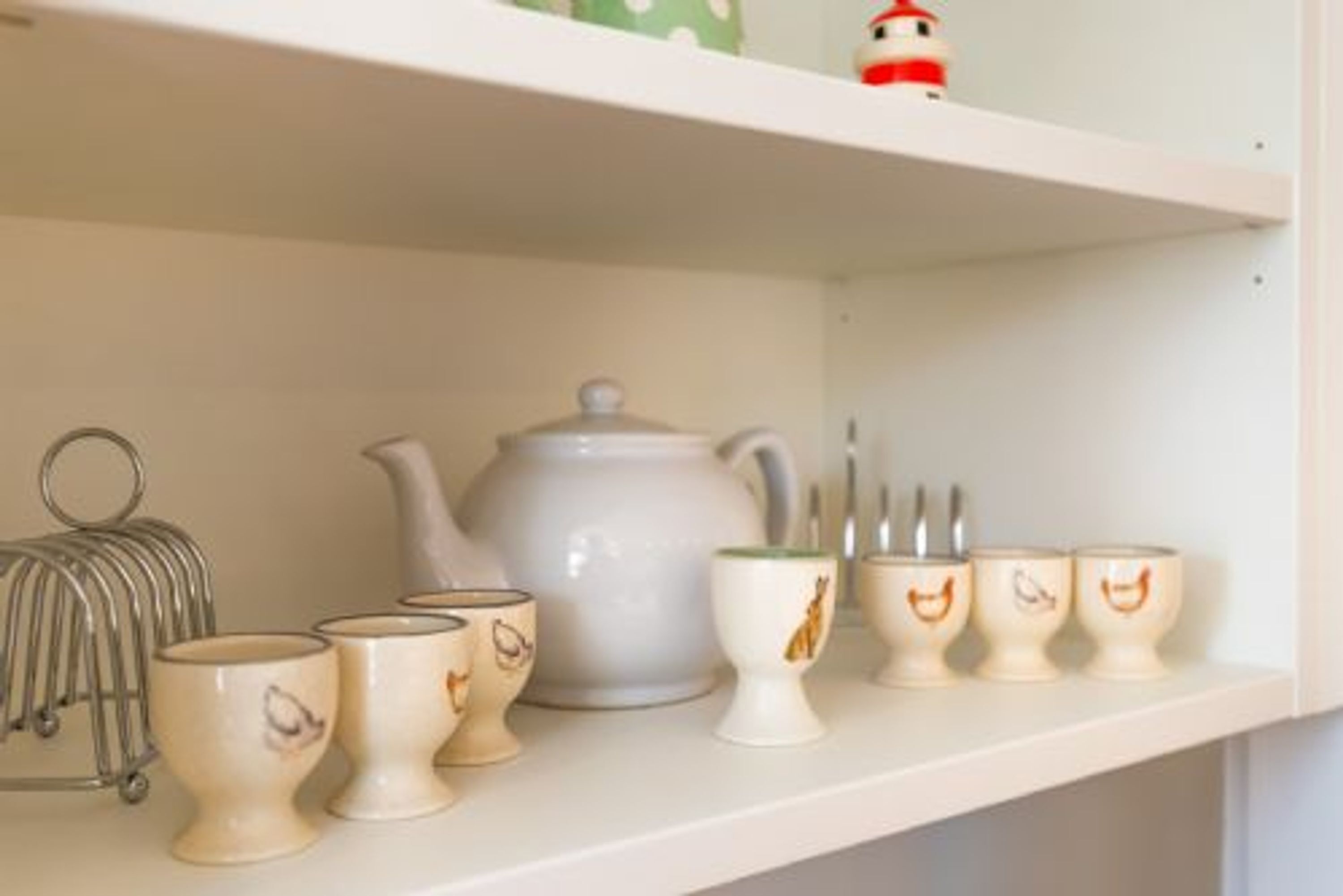 A shelf with a white teapot, several teacups with handles, a metal teapot holder, and a small figurine.