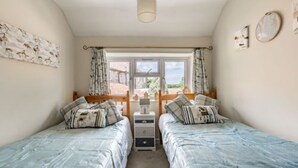 A bedroom with two beds, a window with curtains, a bedside table, and a wall-mounted artwork. - Host Stay Sandpipers (Holt)