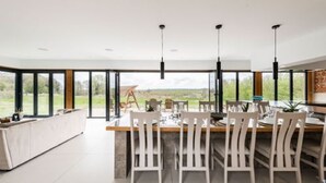 A modern kitchen with a large island, white chairs, and a view of the outdoors through large windows. - Host Stay Willow Barn (Norwich)