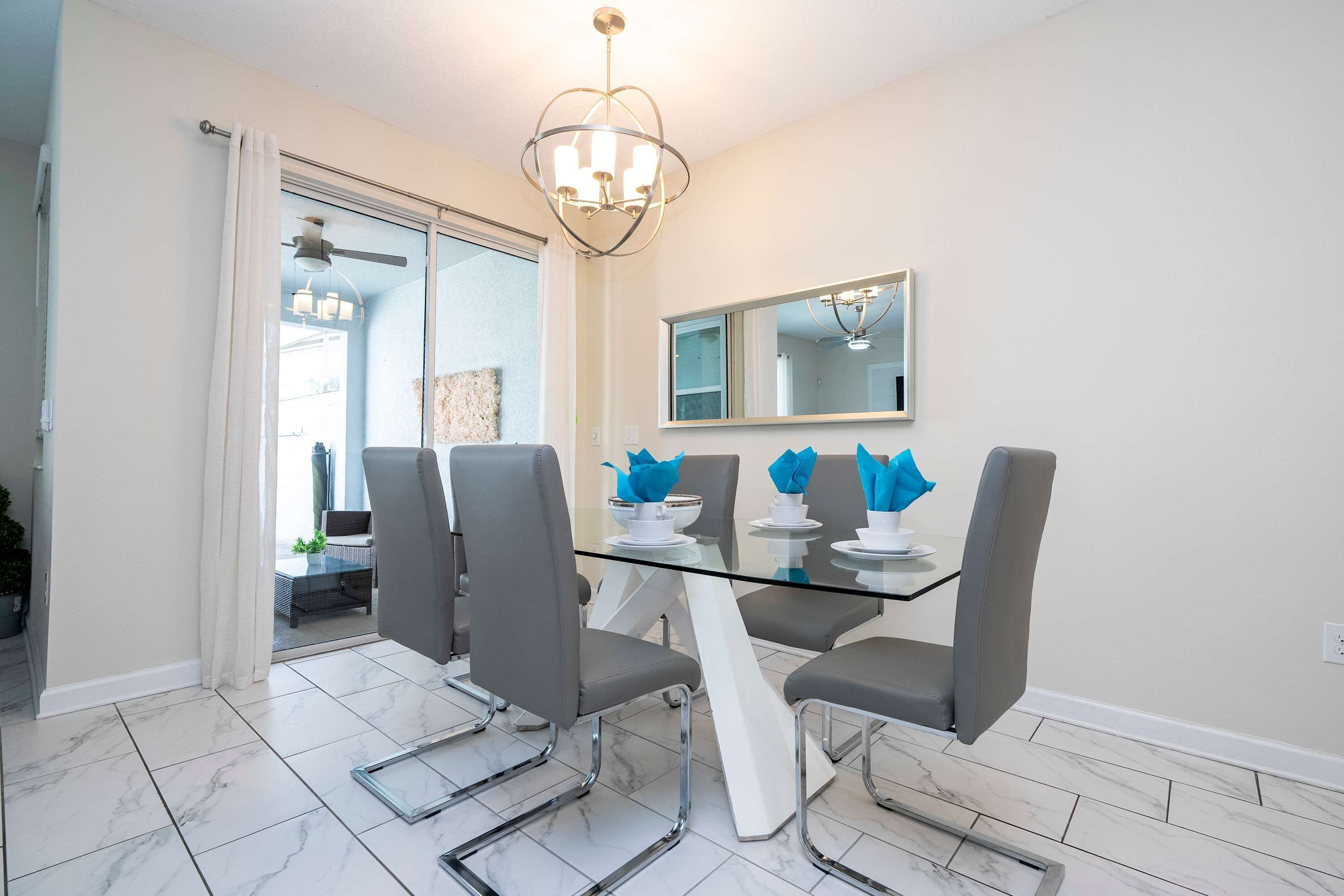 A dining area with a glass table, four grey chairs, a chandelier, and a large mirror on the wall.