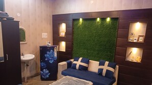 A room with a sofa, a small table, a refrigerator, a sink, and a wall with a green artificial grass panel. - ASHIANA HOME STAY  (Jammu)