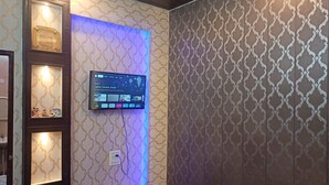 A room with a mounted television displaying a Google homepage, a shelf with decorative items, and patterned wallpaper. - ASHIANA HOME STAY  (Jammu)