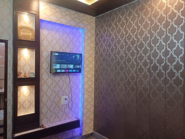 A room with a mounted television displaying a Google homepage, a shelf with decorative items, and patterned wallpaper. - ASHIANA HOME STAY (Jammu)