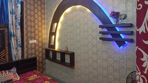 A room with patterned wallpaper, a bed with a colorful quilt, a wall-mounted shelf, and a decorative arch with lighting. - ASHIANA HOME STAY  (Jammu)