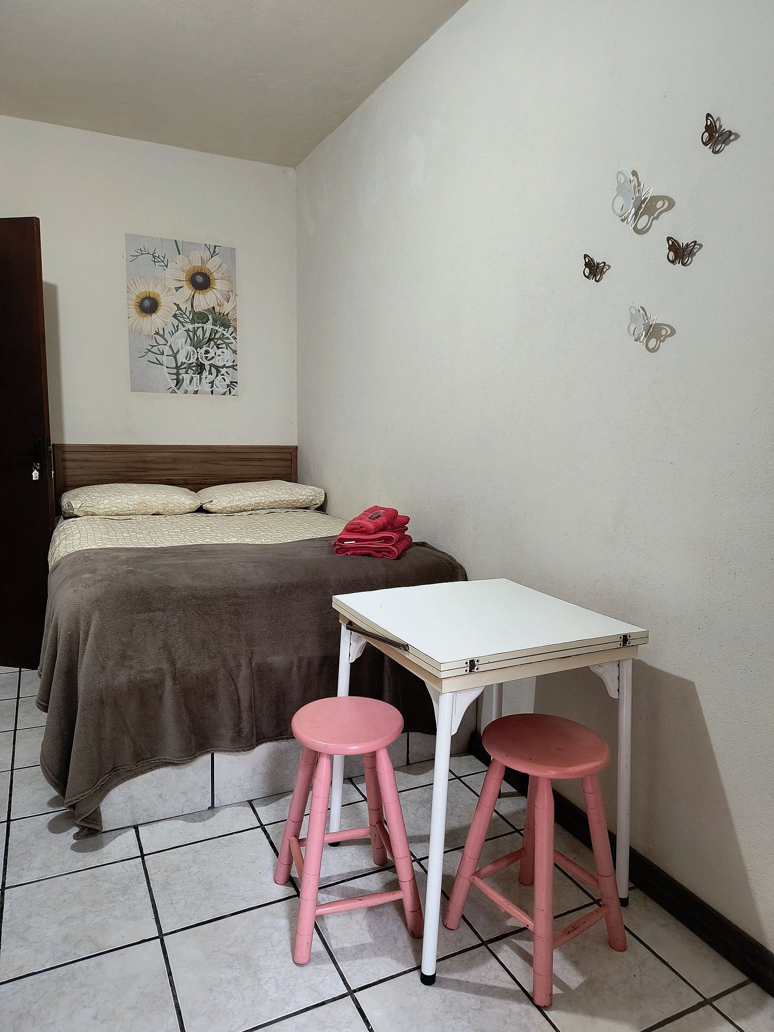 A bedroom with a bed, a small table, two pink stools, a red bag on the bed, and butterfly wall decals.