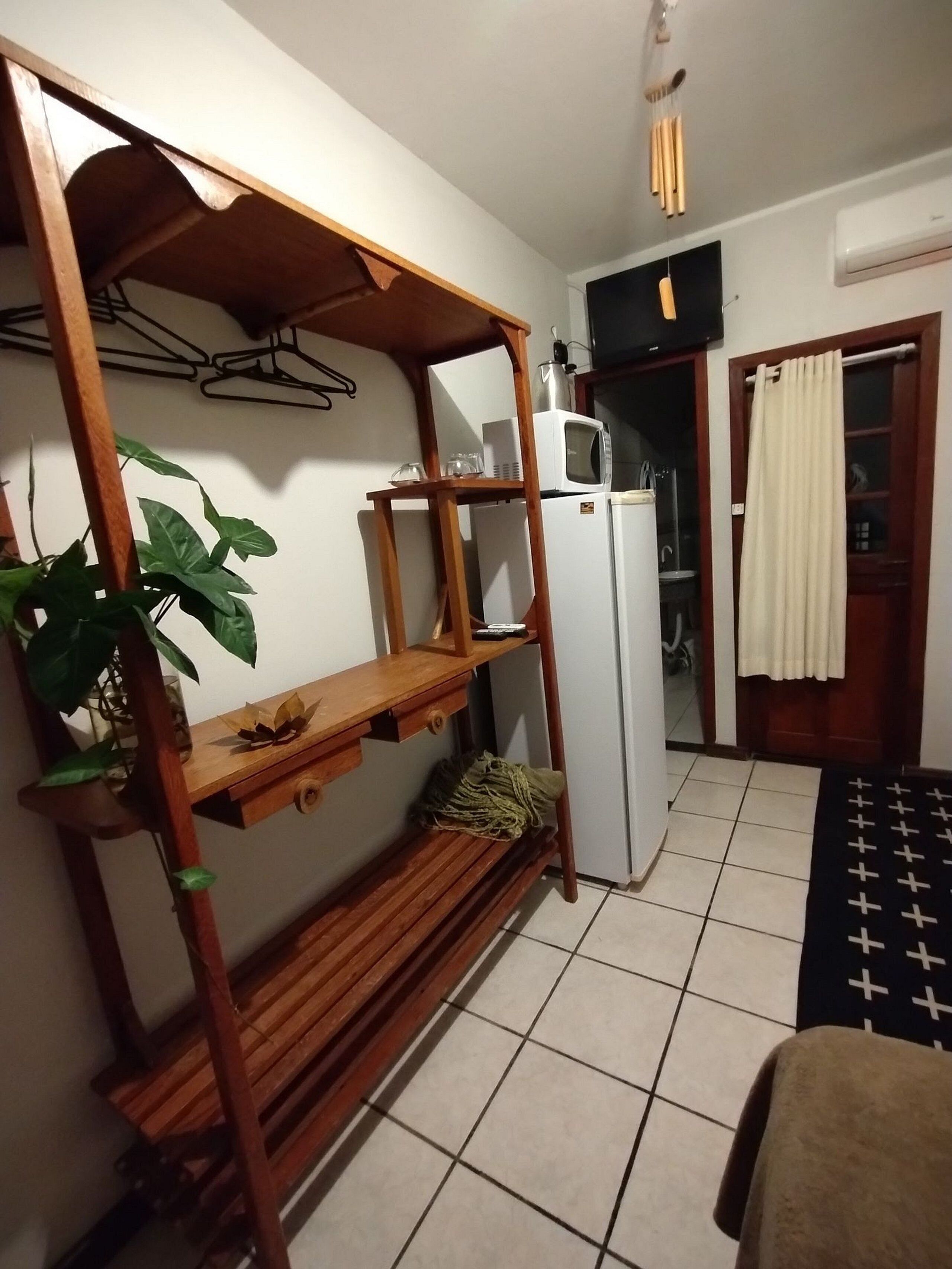 A compact kitchen with a wooden shelf unit, a refrigerator, a microwave, and a potted plant.