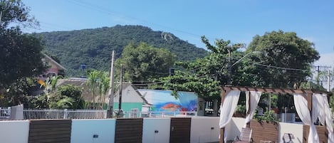 Piscina retangular com deck de madeira, cercada por uma cerca branca e uma pérgola de madeira com cortinas brancas.
