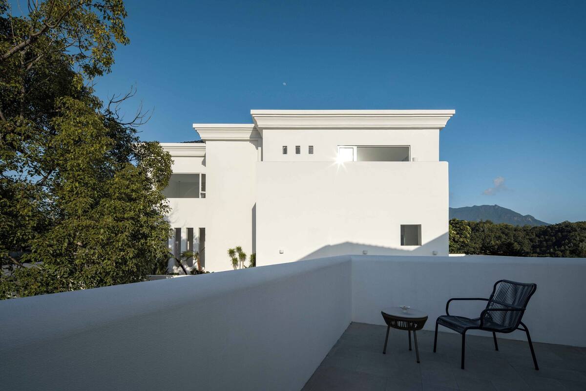 En moderne hvid bygning med fladt tag, en balkon med to stole og udsigt til et bjerglandskab.