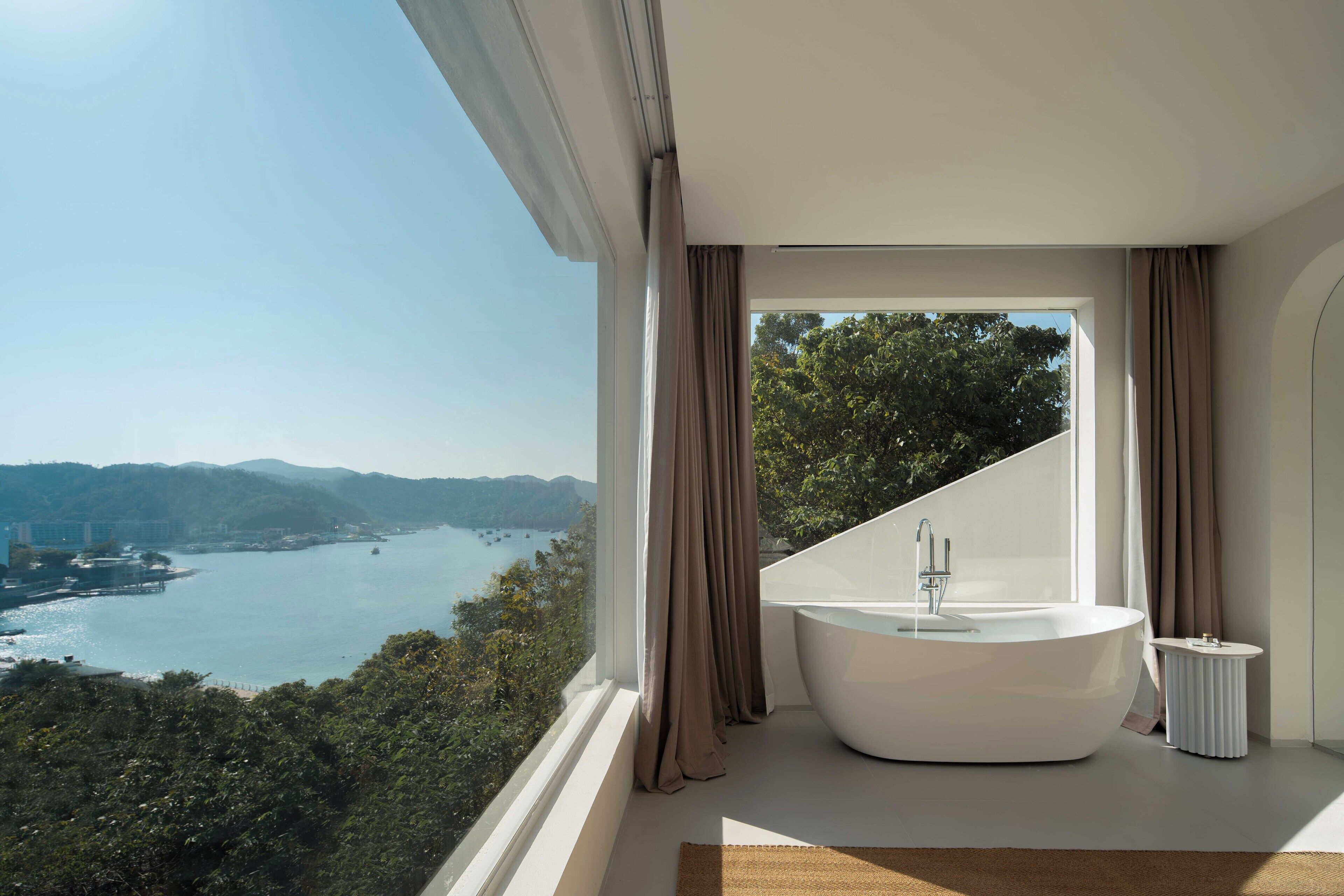 A modern bathroom with a freestanding bathtub, large windows, and a view of a lake and mountains.