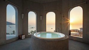 Une salle de bain moderne avec une baignoire sur pieds, de grandes fenêtres en arc de cercle et une vue sur la mer au coucher du soleil.