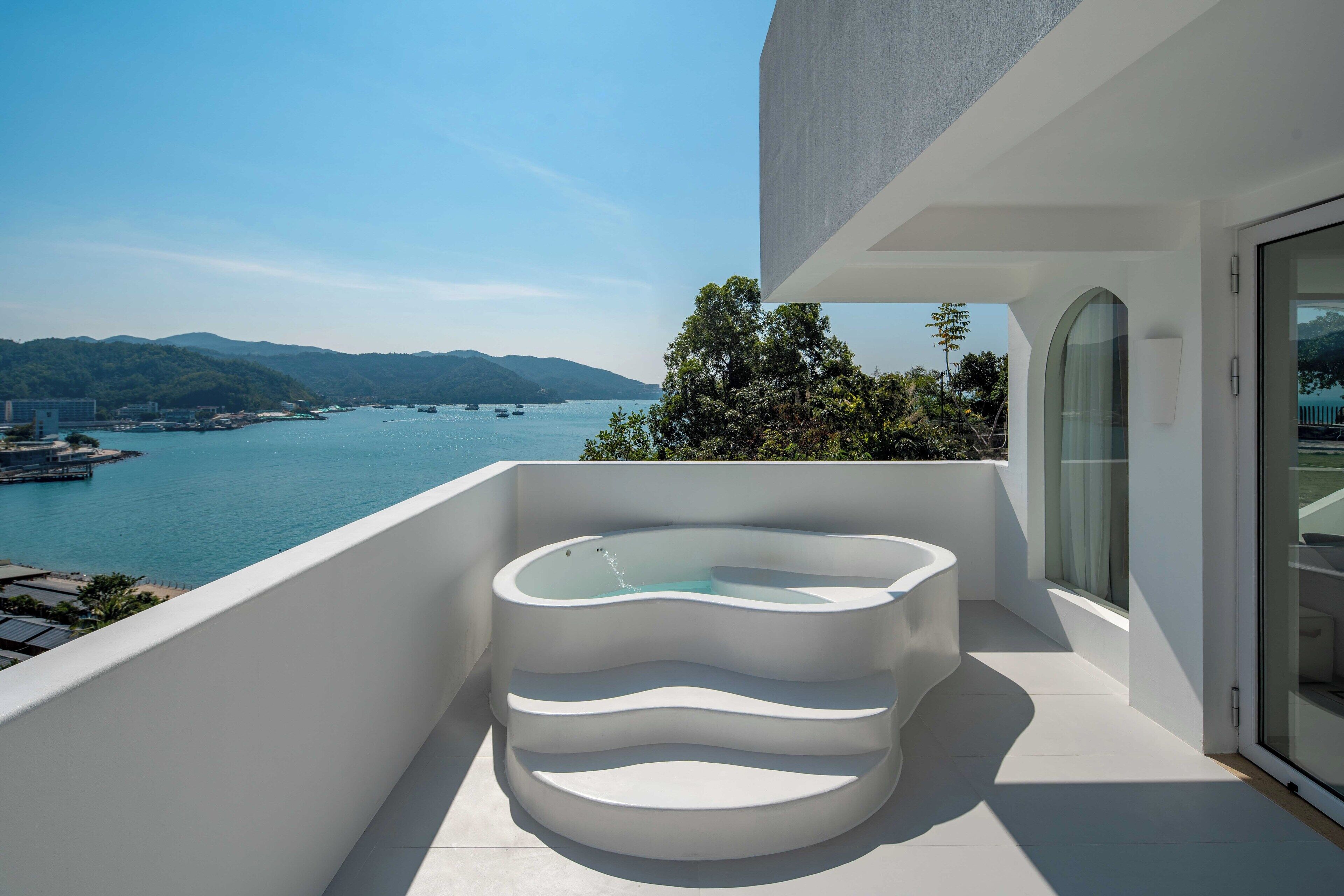 A modern balcony with a unique, multi-tiered hot tub overlooking a coastal bay with boats and hills.
