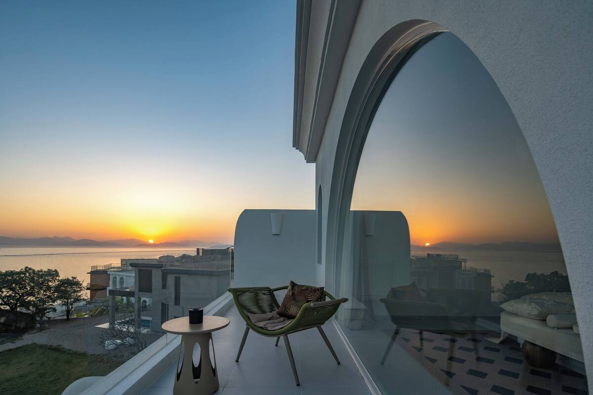 En balkon med udsigt til solnedgangen over vandet, en grøn stol og et lille bord.
