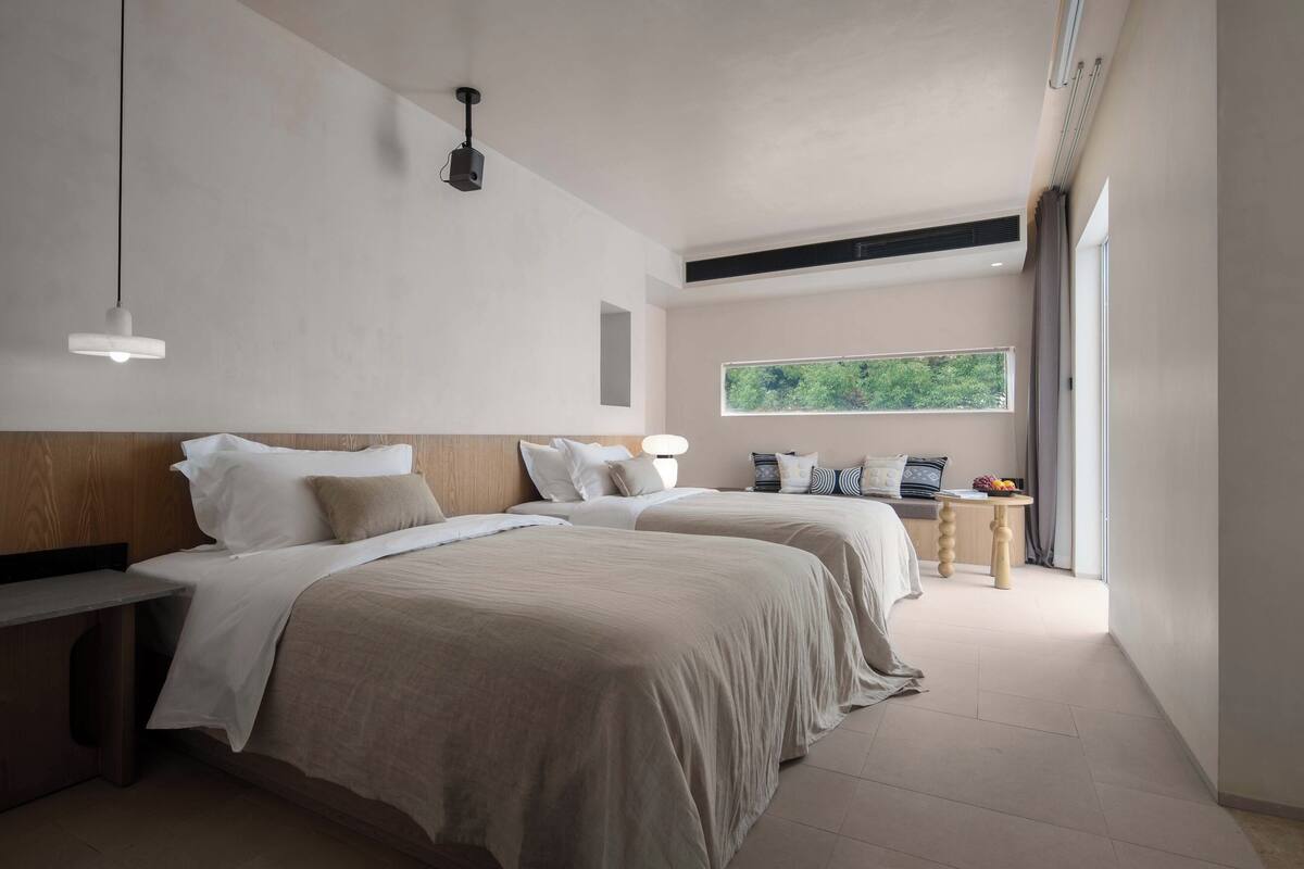 Et moderne hotelværelse med to senge, et lille bord og et stort vindue med udsigt til grønne områder.
