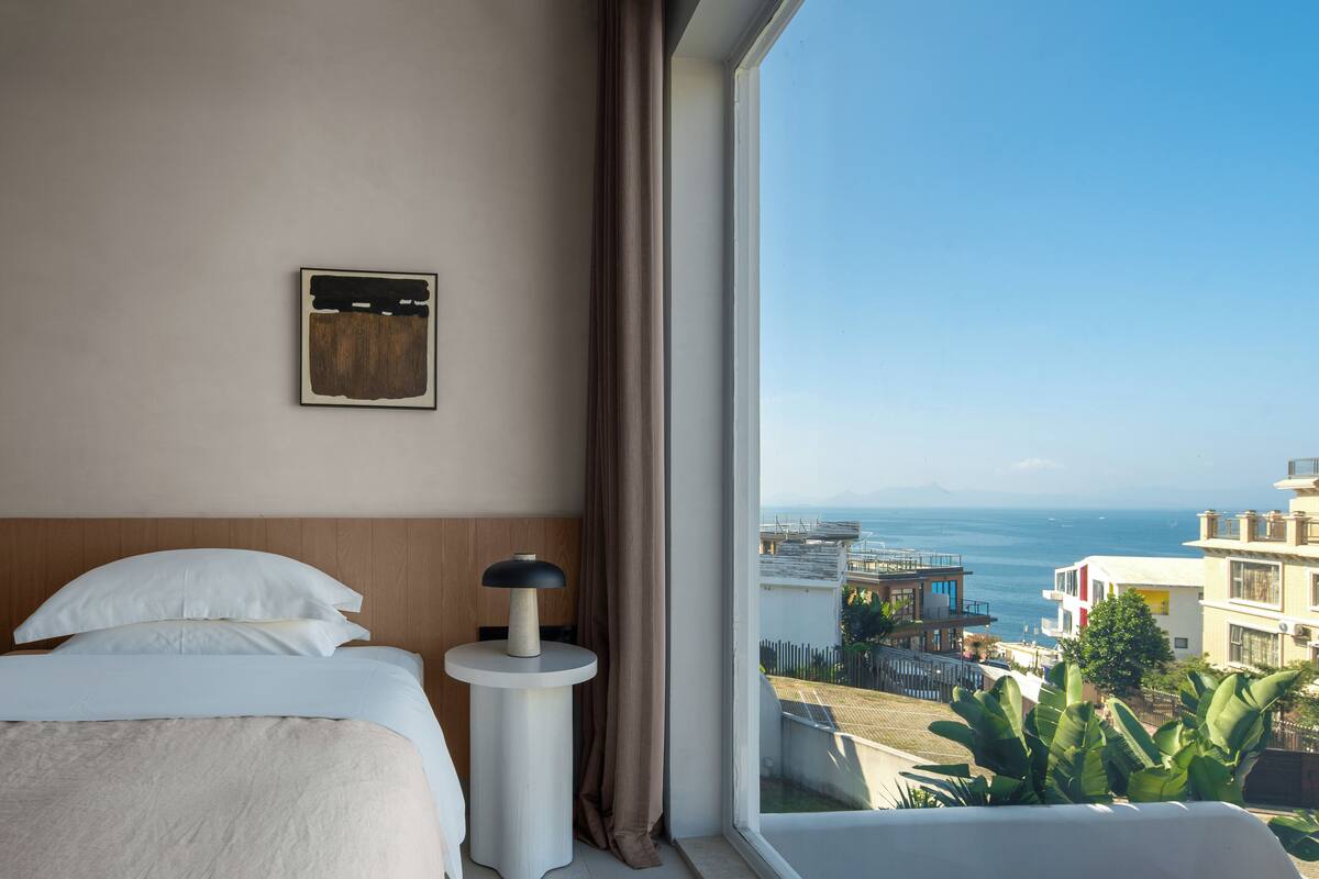 Et hotelværelse med en seng, et sengebord, en lampe, et vindue med udsigt til havet og bygninger.
