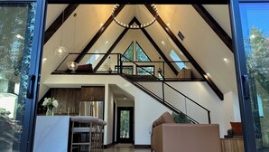 A spacious loft with a high ceiling, a staircase leading to an upper level, a kitchen area, and a seating area with a sofa. - Everwoods Aframe - Modern Luxury, Cozy Mountain Retreat (McCall)