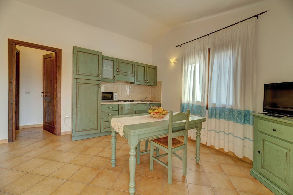 Une cuisine de taille réduite avec des meubles de couleur verte, une table à manger avec des chaises, un panier de fruits et un téléviseur.