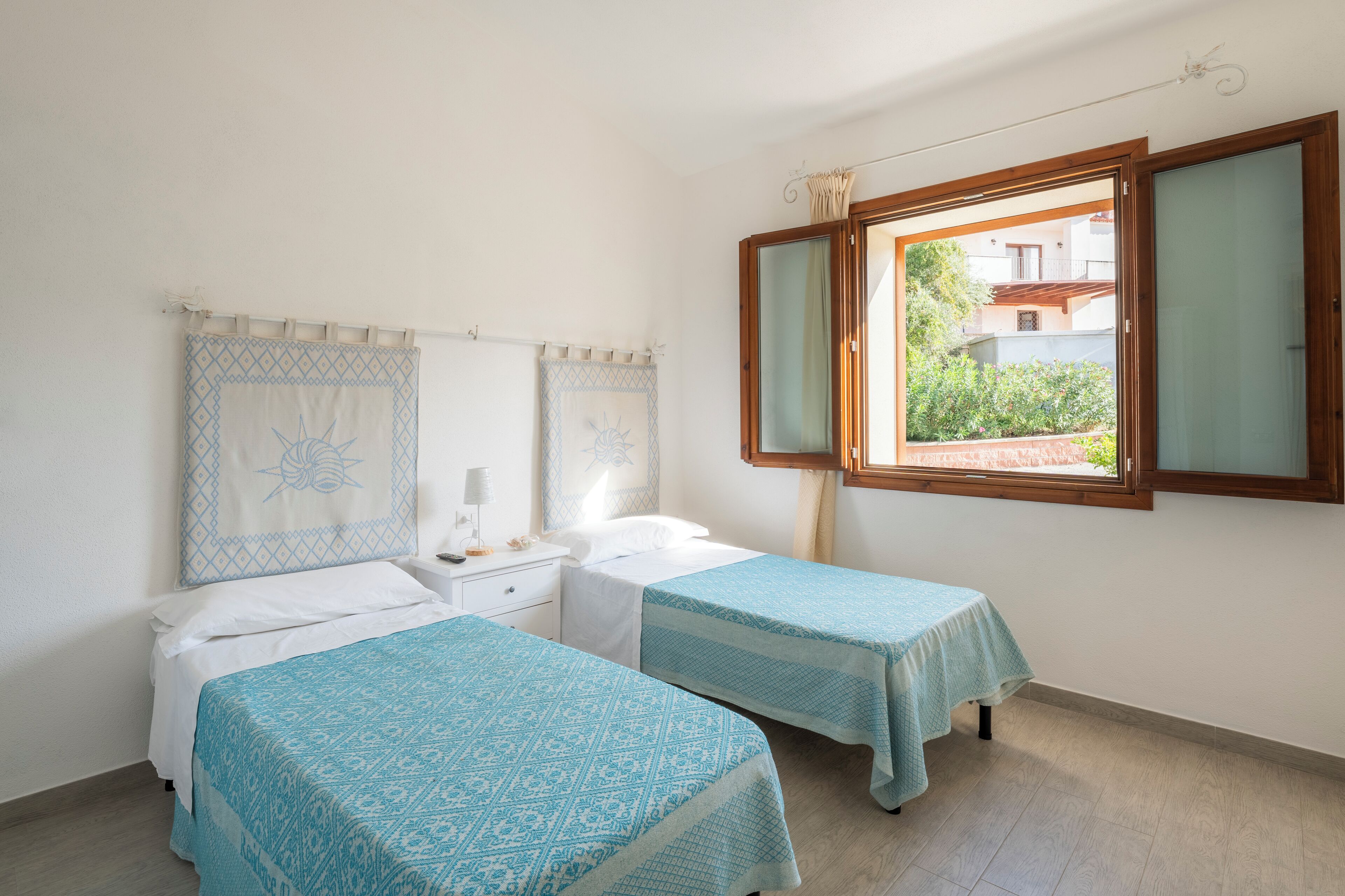 A bedroom with two beds, a nightstand, a mirror, and a window with a view of buildings and greenery.