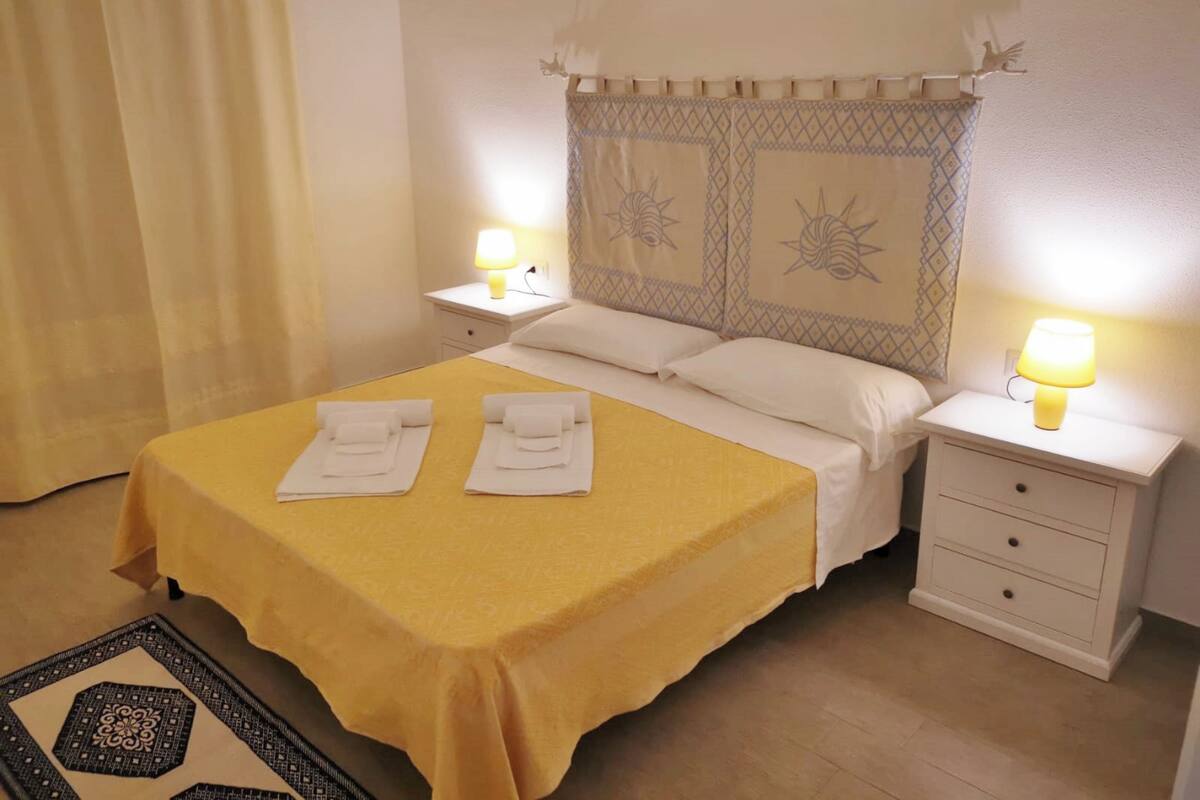 Une chambre d’hôtel avec une couvre-lit jaune, deux serviettes blanches, une tête de lit décorative, deux lampes de chevet et une table de chevet avec des tiroirs.