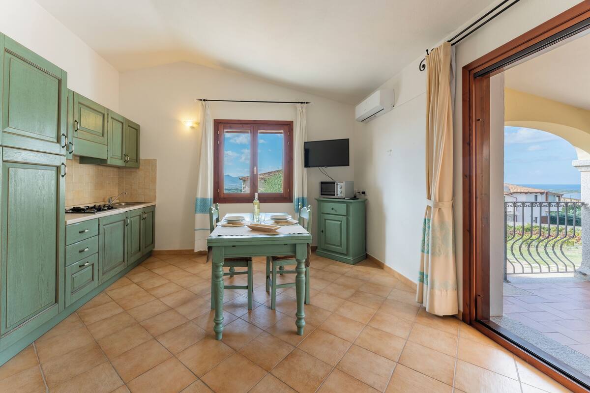 Une cuisine avec des meubles de couleur verte, une table à manger, un téléviseur et un balcon avec vue.