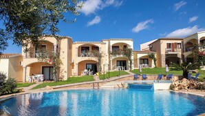 A swimming pool surrounded by a building with balconies, outdoor seating, and a clear blue sky. - I Giardini di Badus (Badesi)