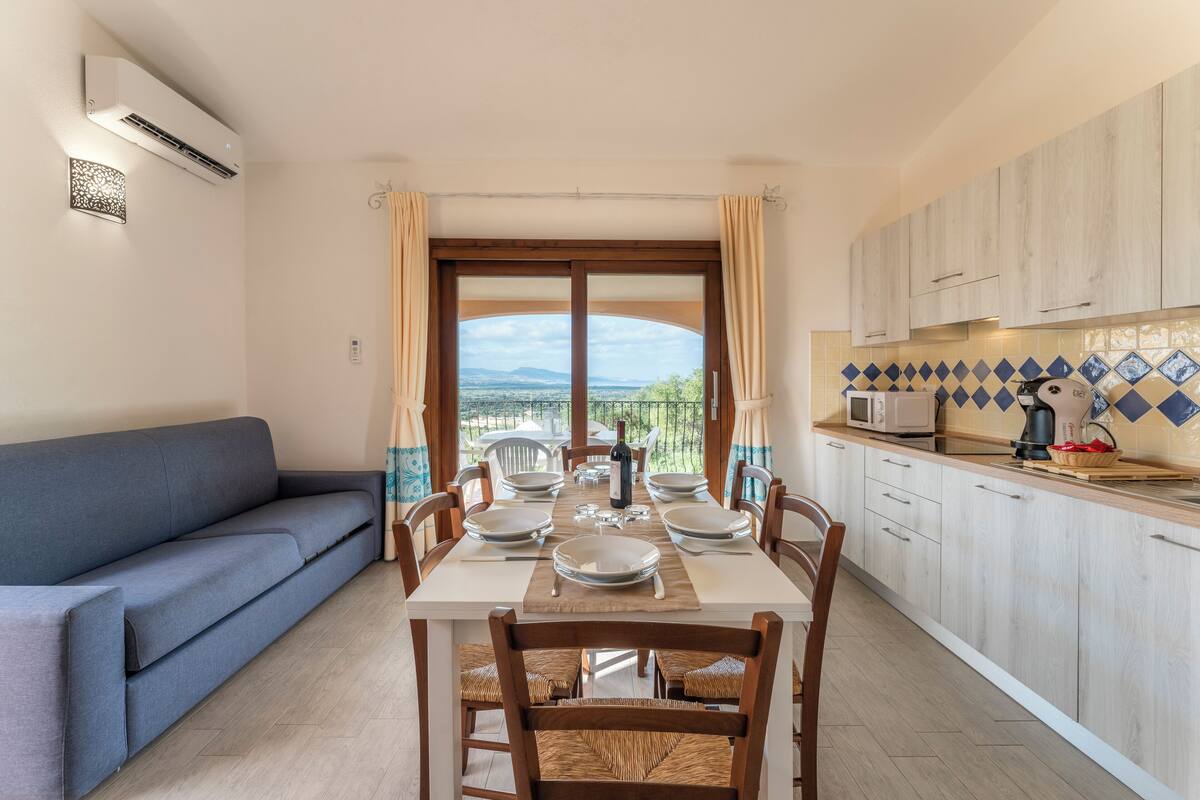 Un coin repas avec une table dressée pour six personnes, un canapé et une cuisine dotée de meubles en bois et d’un plan de travail carrelé.