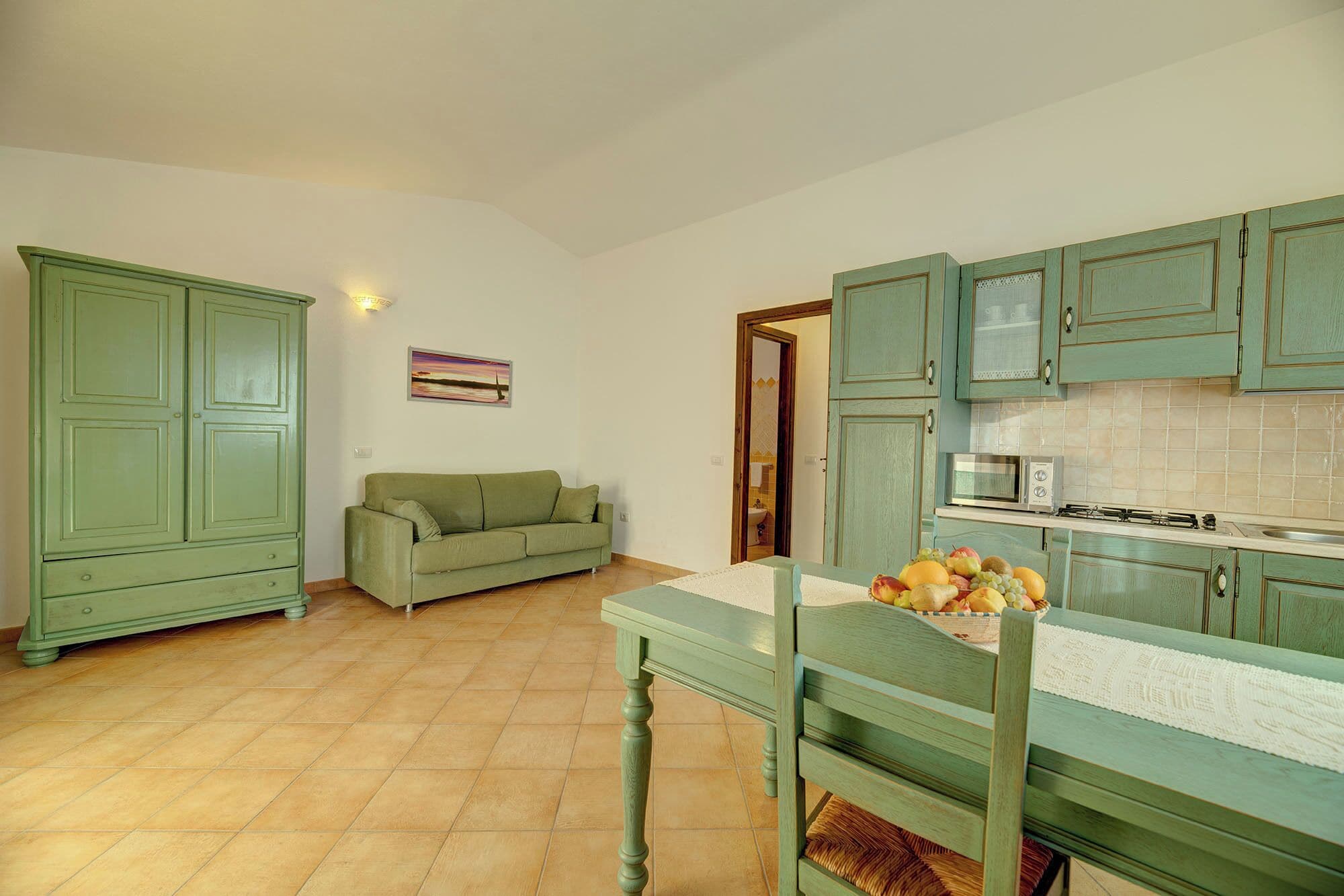 A compact kitchen with green cabinets, a dining table with a fruit basket, and a green sofa in a small living area.