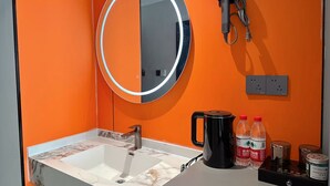 A bathroom with an orange wall, a round mirror with a light, a hairdryer, a sink with a marble countertop, and toiletries on the counter. - Qin Mengli Hotel (Xianyang)