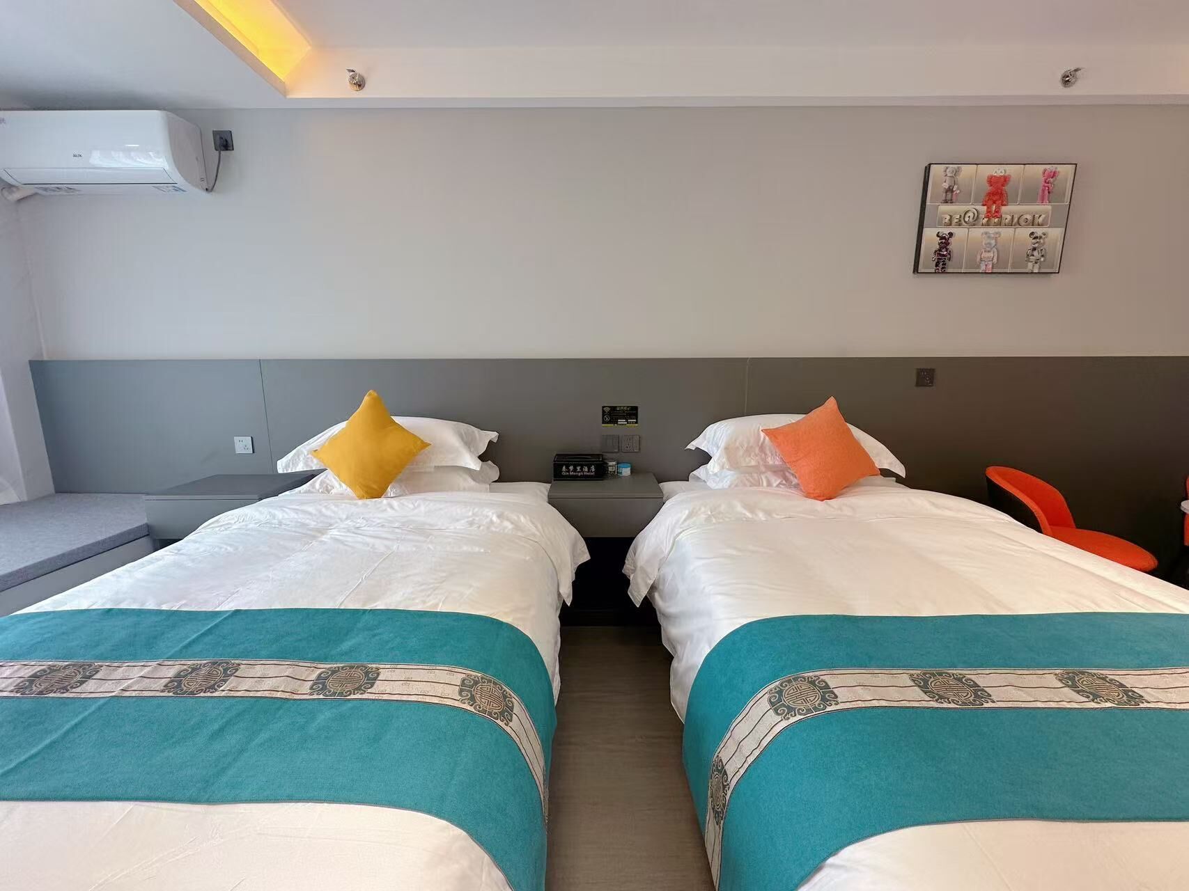 A hotel room with two beds, each with a teal bedspread and white bedding. There is a small table with a lamp and a framed picture on the wall.