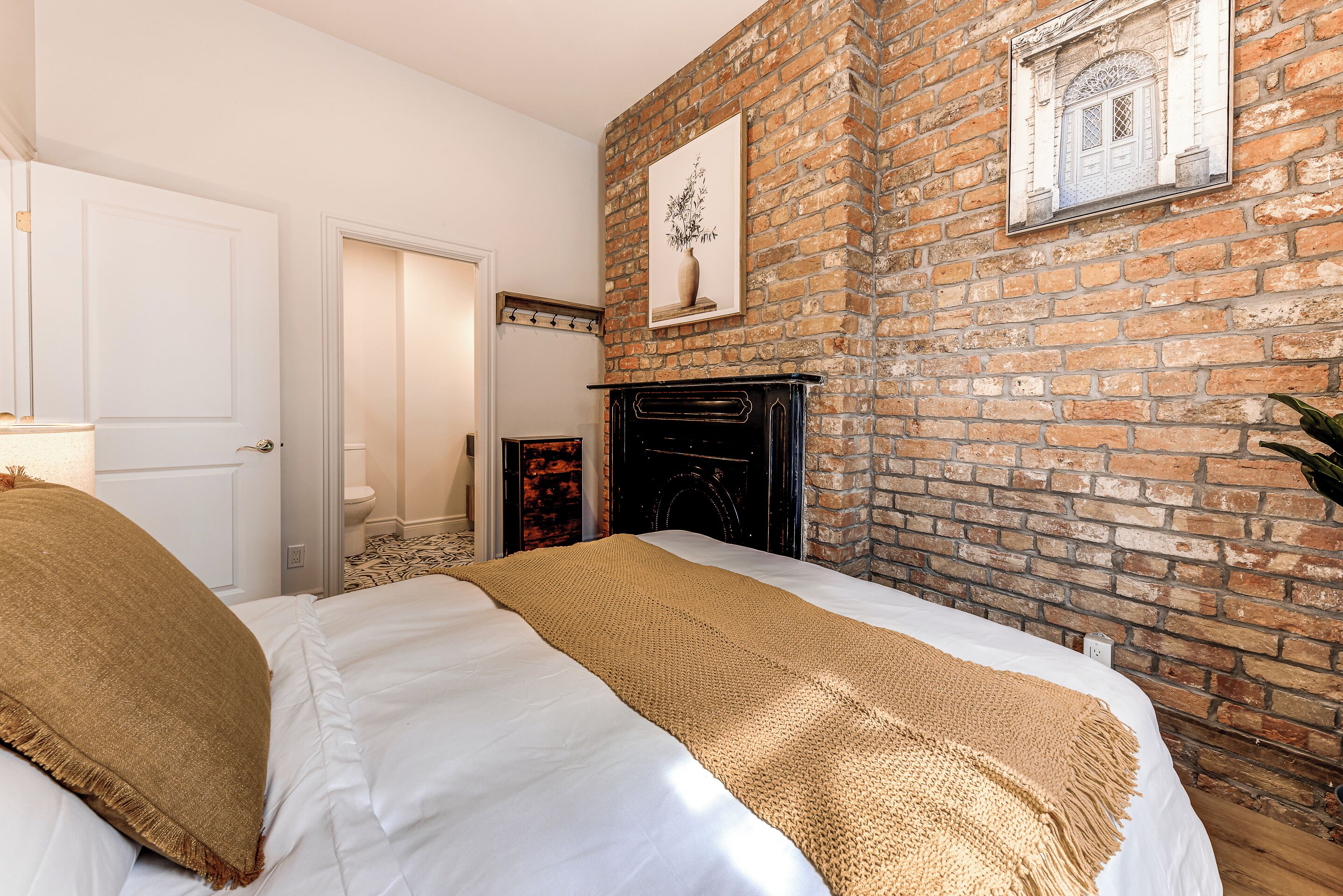 A bedroom with a brick wall, a fireplace, and a bed with white bedding and a beige throw blanket.