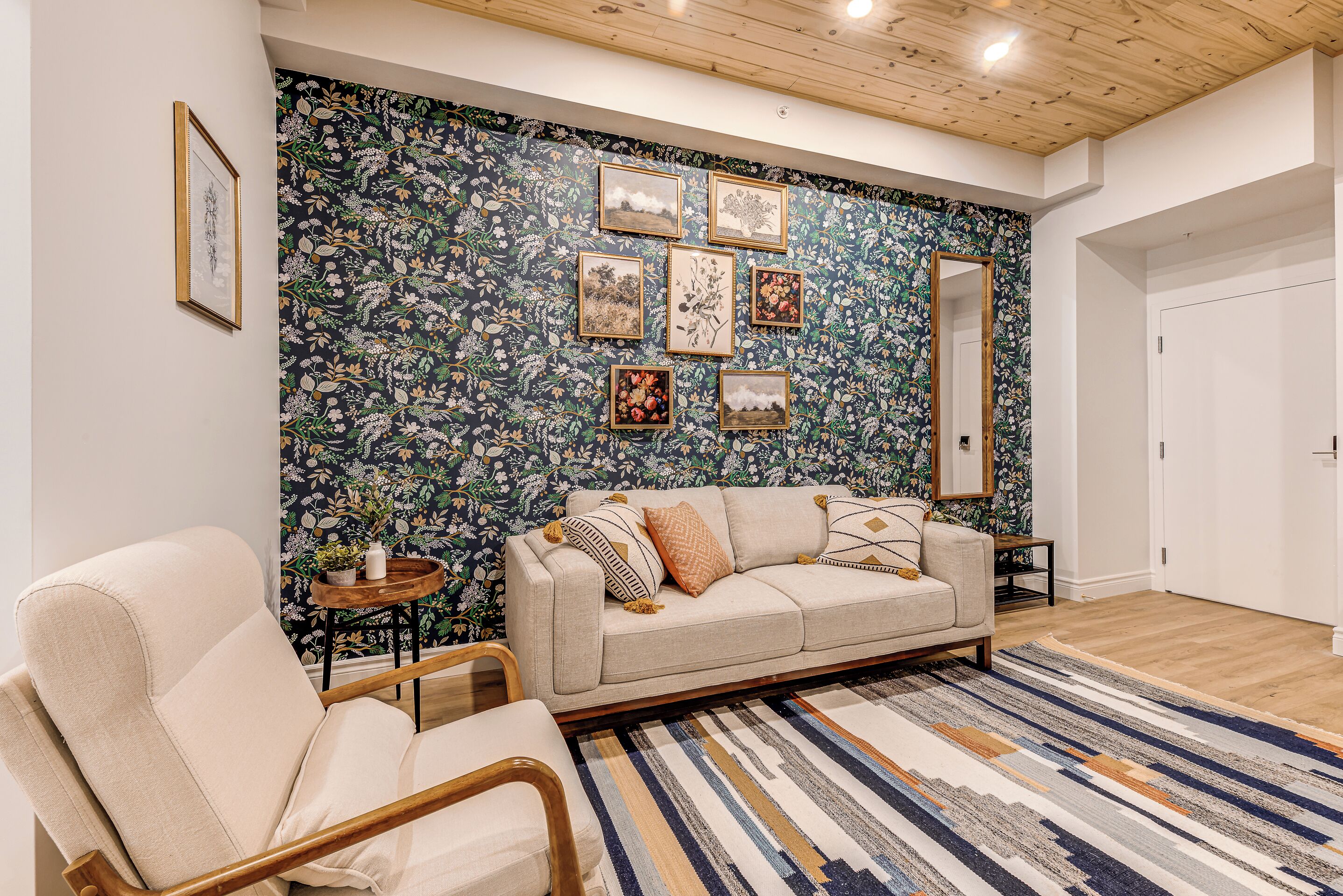 A living room with a floral wallpaper, a beige sofa, a striped rug, and framed pictures on the wall.
