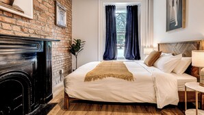 A bedroom with a brick wall, a fireplace, a large bed with white bedding, and a window with curtains. - Historic Two Bedroom Facing Parliament (Québec)