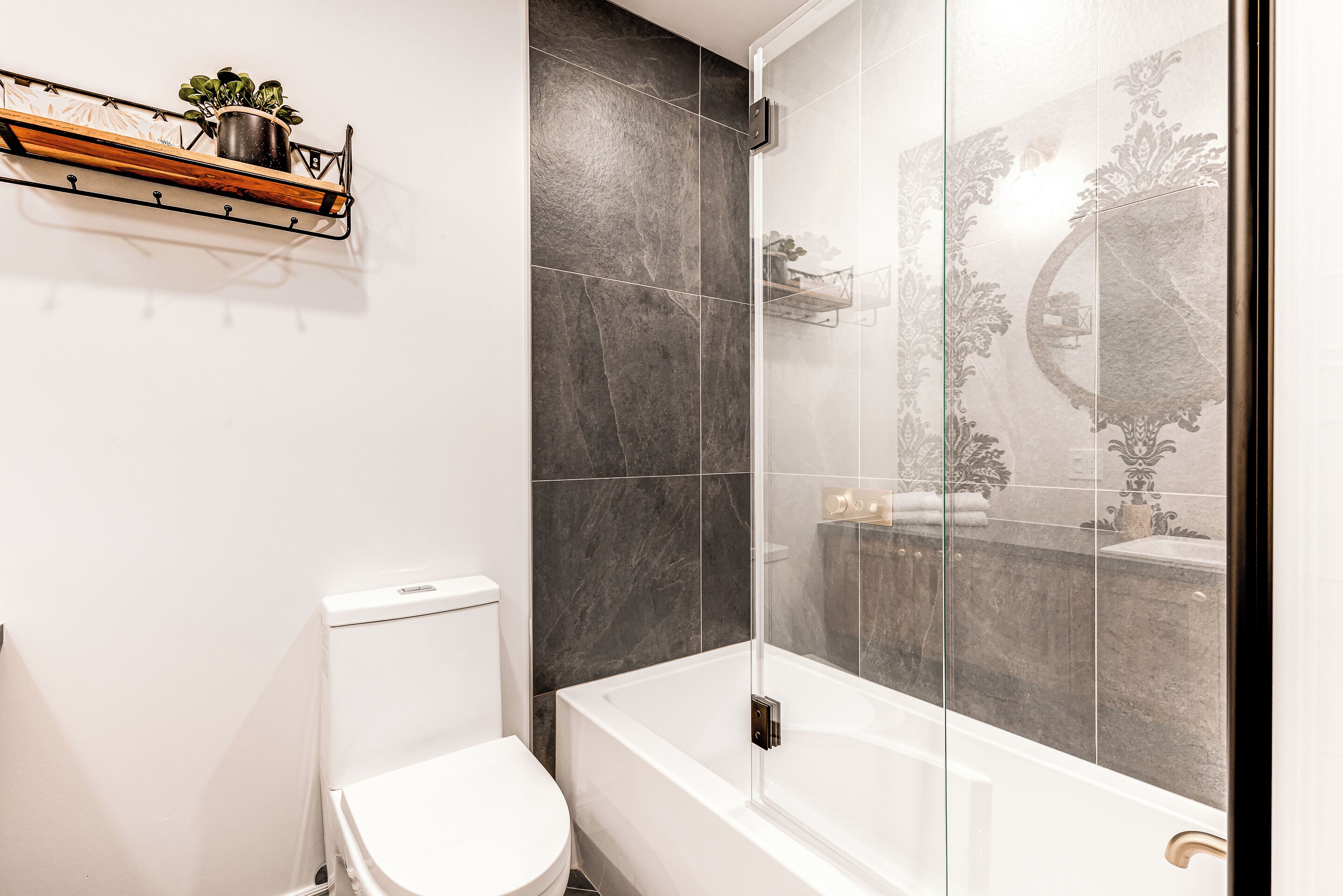 Un baño con inodoro blanco, bañera con cerramiento de ducha de vidrio y estante con planta en maceta.