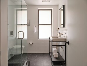 A modern bathroom with a glass shower enclosure, a toilet, a sink with a mirror, and a window providing natural light. - Mint House Washington DC Downtown by Kasa (Washington)