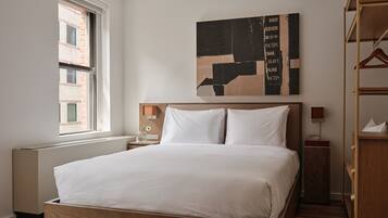 A modern hotel room with a large bed, a wooden headboard, a nightstand with a lamp, a window with a view of buildings, and a wall-mounted abstract artwork.