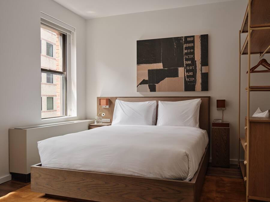 A modern hotel room with a large bed, a wooden headboard, a nightstand with a lamp, a window with a view of buildings, and a wall-mounted abstract artwork.