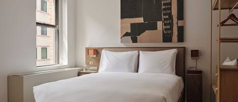 Camera d'albergo moderna con un letto grande, una testiera in legno, un comodino con una lampada, un'opera d'arte a parete e una finestra con vista su un edificio in mattoni.