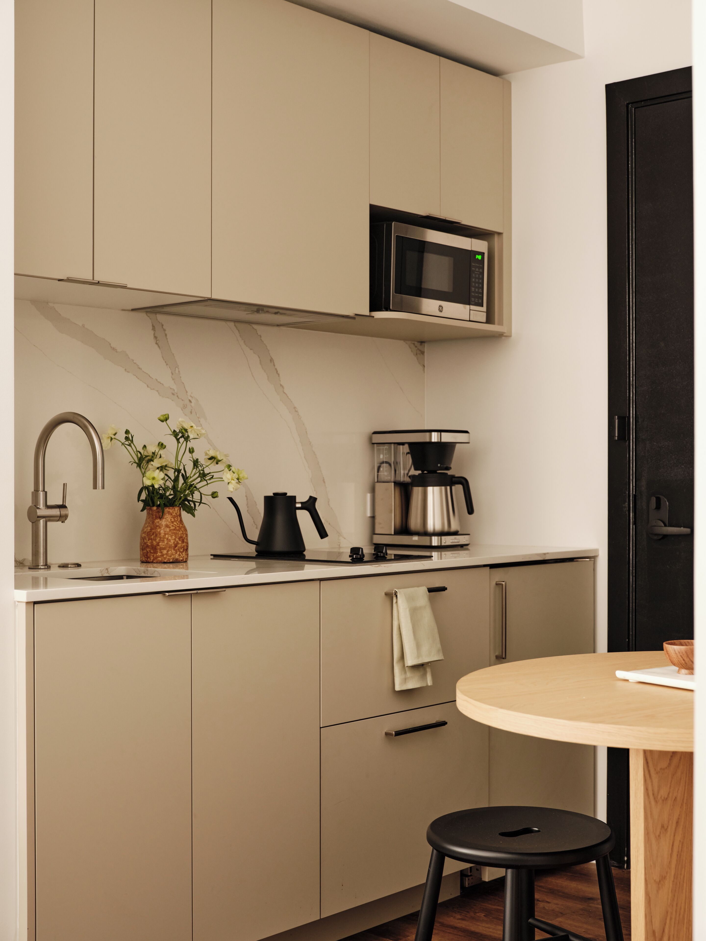 A modern kitchen with a microwave, kettle, and coffee maker. There is a sink with a faucet, a vase with flowers, and a wooden table with a stool.