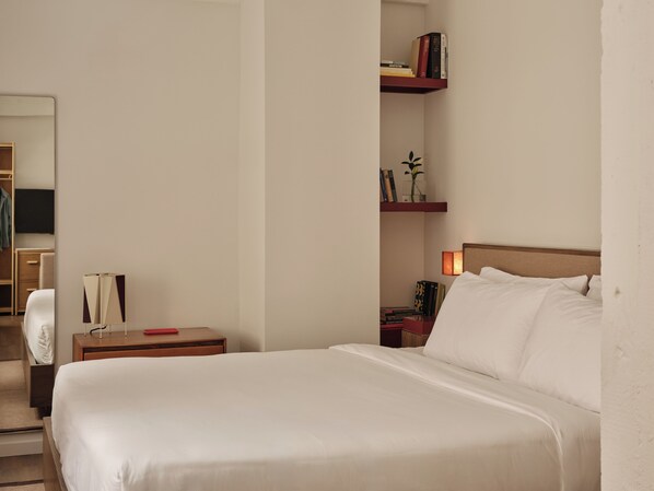 A hotel room with a large bed, a bedside table with a lamp, a nightstand with books and a vase, and a mirror on the wall. - Mint House Washington DC Downtown by Kasa (Washington)