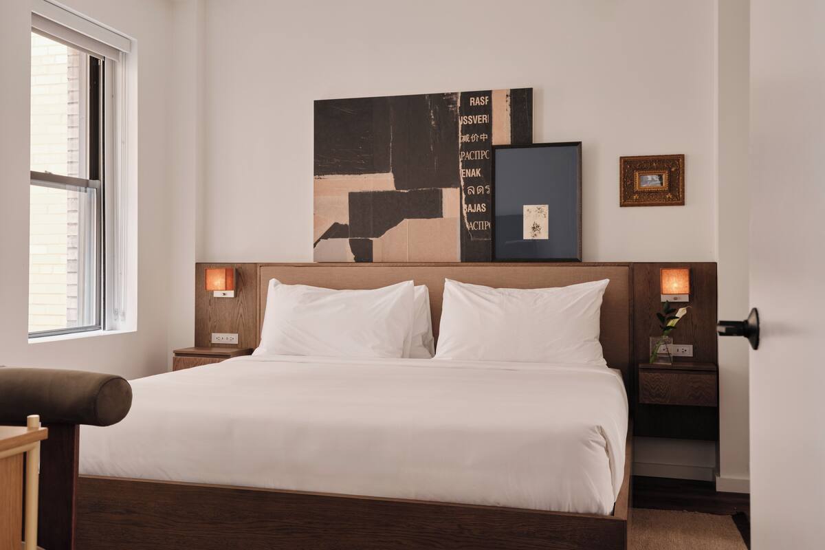Une chambre d’hôtel avec un lit en bois, du linge de lit blanc, un grand tableau abstrait accroché au mur et deux tables de chevet avec des lampes.