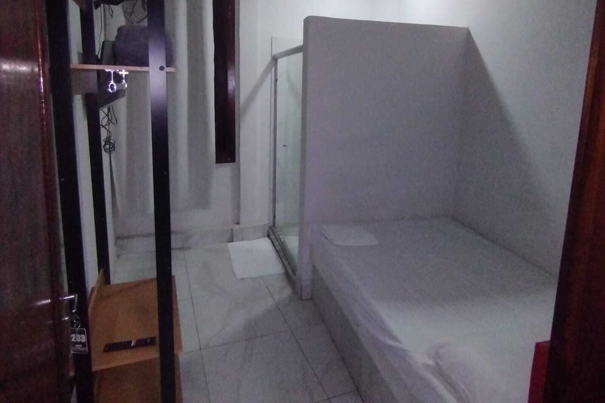 A small, dimly lit room with a single bed, a small table, and a door leading to another room.