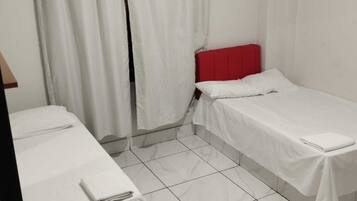 Quarto pequeno e limpo, com duas camas de solteiro, cortinas brancas e chão revestido de azulejos.