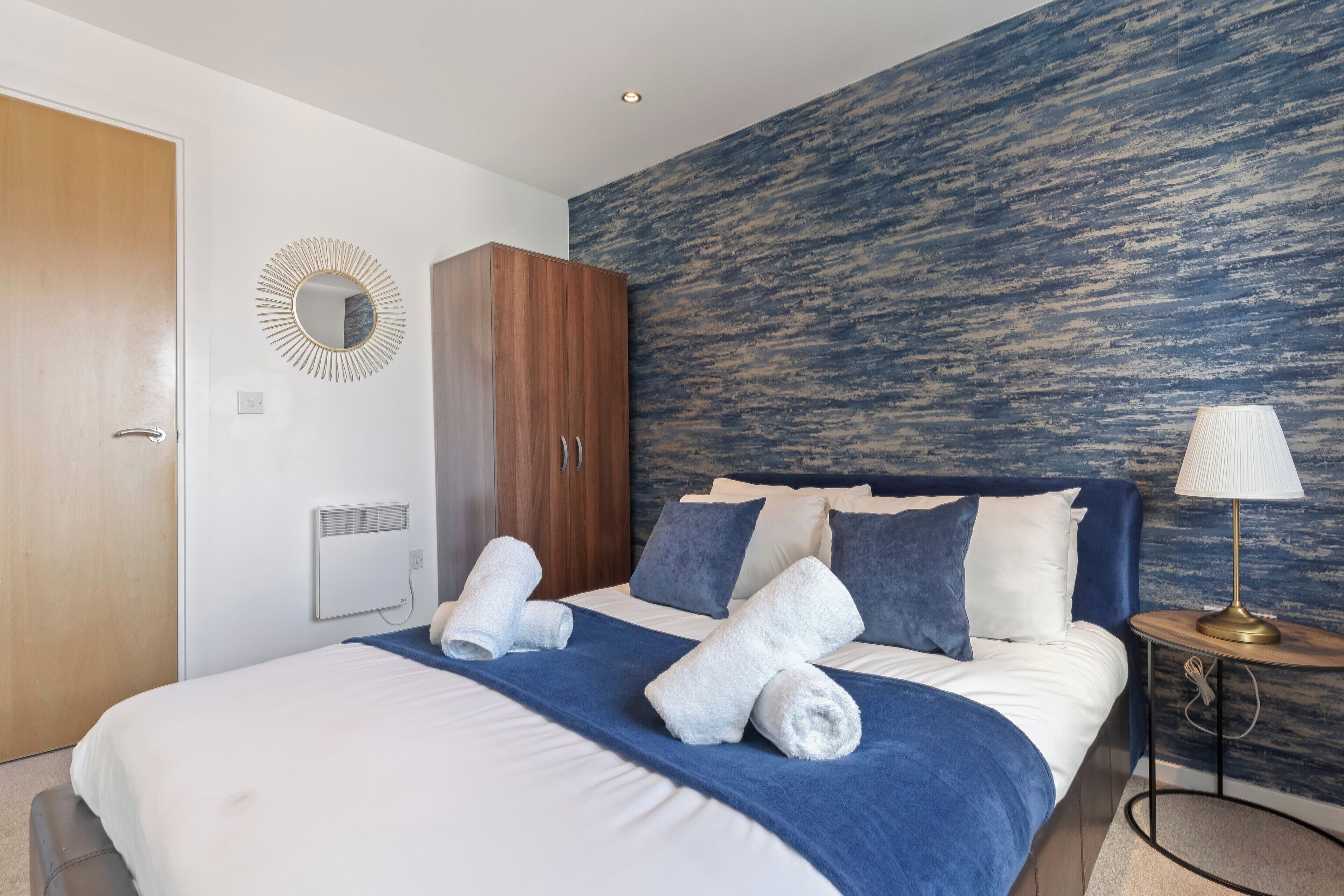 A modern hotel room with a large bed, a wooden wardrobe, a round mirror, and a textured wall.