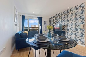 Un coin repas avec une table ronde dressée pour quatre personnes, des canapés bleus et une vue sur la ville par une fenêtre ornée de rideaux bleus.