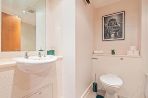 Un bany modern amb un lavabo blanc, un mirall i un vàter. Hi ha un quadre a la paret i una petita planta a l'estant.