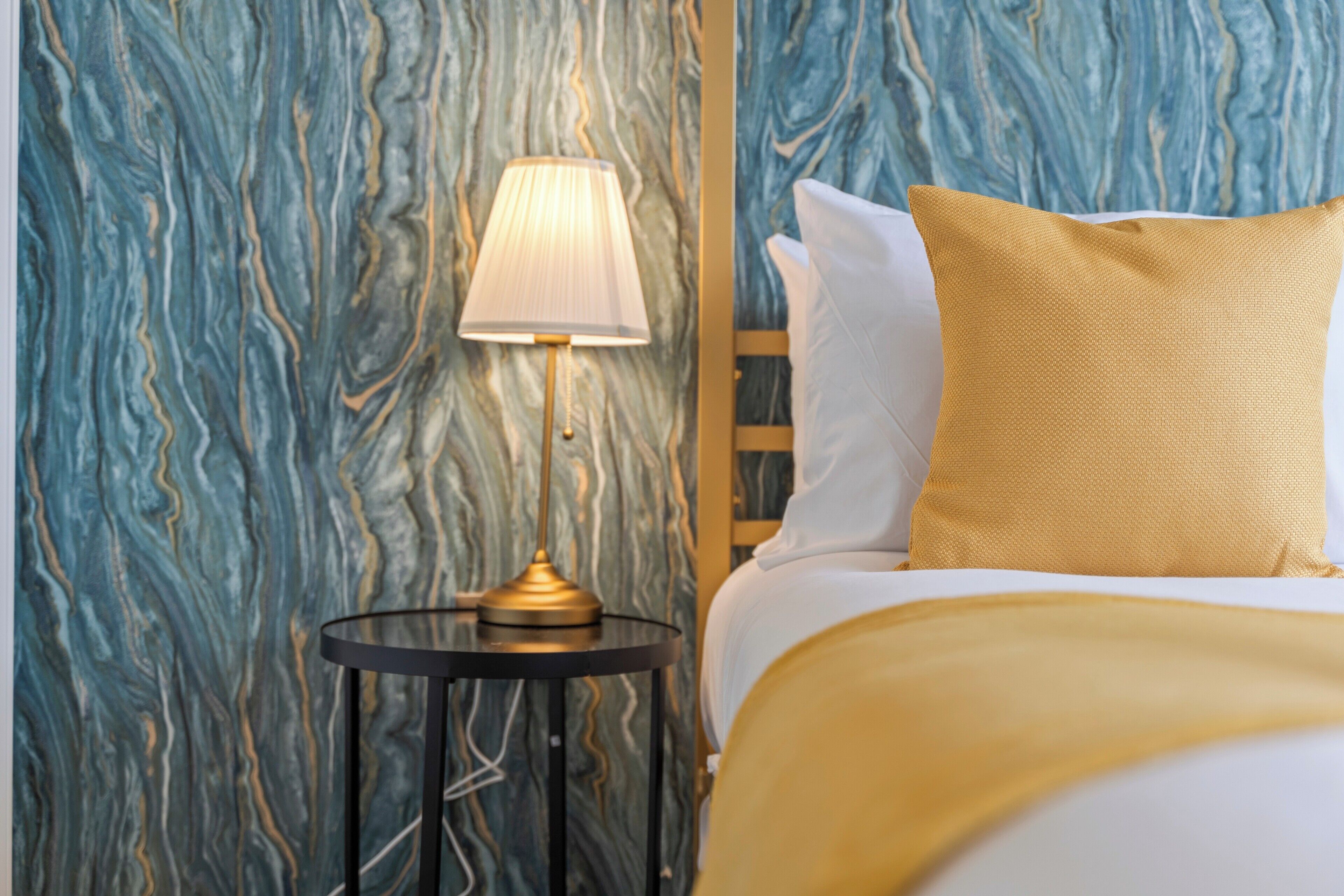 Ein Bett mit weißen Leinen und einem goldenen Kopfkissen, ein runder Nachttisch in Schwarz mit einer Lampe und eine Wand mit Marmoroptik.