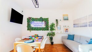 Ein Essbereich mit Tisch für vier Personen, einem wandmontierten Flachbildfernseher, einer grünen Wanddekoration aus Blättern und einem grauen Sofa mit blauen Kissen.