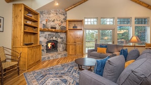 Una sala de estar acogedora con chimenea de piedra, estanterías de madera, un sofá gris con cojines azules y amarillos, y grandes ventanales que ofrecen vistas al exterior.
